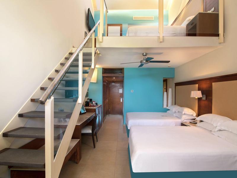 Moderne hotelkamer met tweepersoonsbedden, bureau, trap naar loft en lichtblauwe muur.