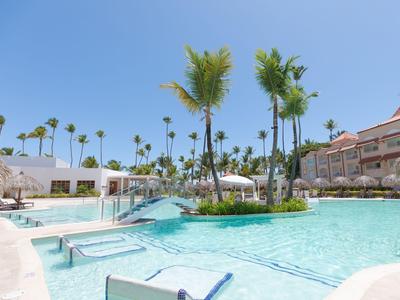 Großer Hotelpool mit Palmen, Sonnenschirmen und Liegestühlen bei klarem Himmel.