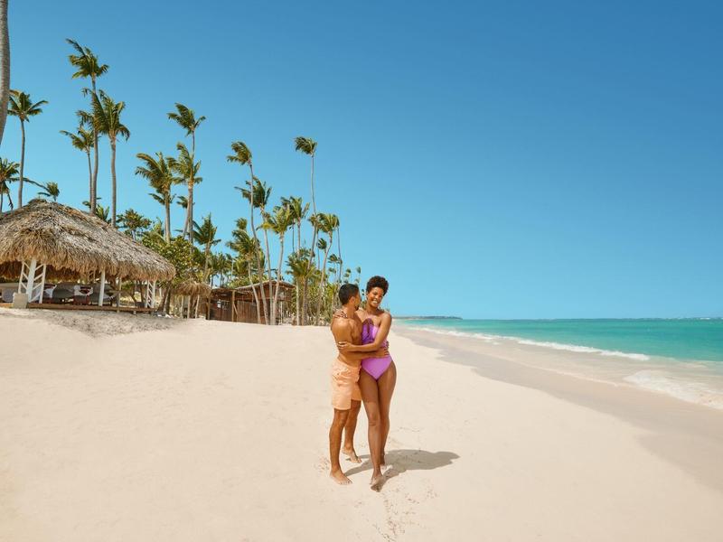 Paar am tropischen Strand mit Palmen und klarem Himmel neben türkisfarbenem Meer.