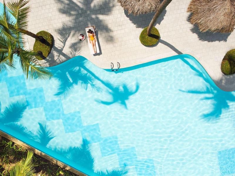 Pool mit klarem blauem Wasser, umgeben von Palmen und Sonnenschirmen, mit Schatten darauf