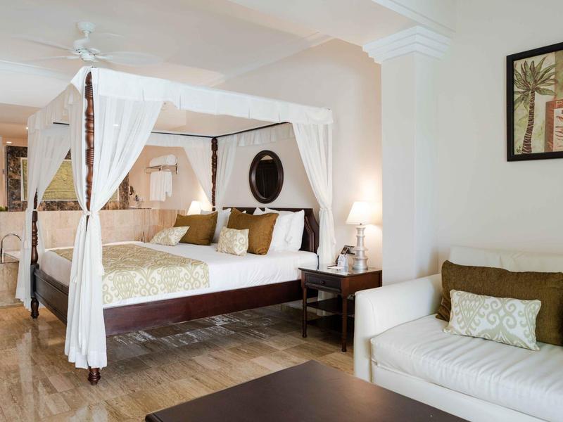 Elegantes Hotelzimmer mit Himmelbett, Sofa, Lampen und Wanddekoration in hellen Farben.