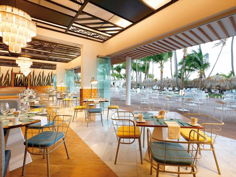 Helles Restaurant mit modernen Möbeln und großer Glasfront zur Terrasse mit Blick auf Palmen.