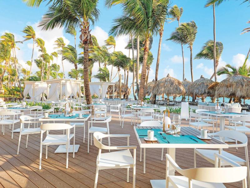 Freiluftrestaurant mit weißen Stühlen und Tischen unter Palmen bei Sonnenlicht.