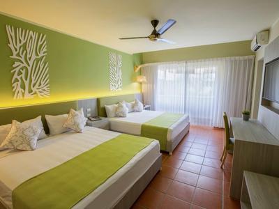 Hellgrünes Hotelzimmer mit zwei großen Betten, weißer Wanddekoration und Terrakottafliesen.