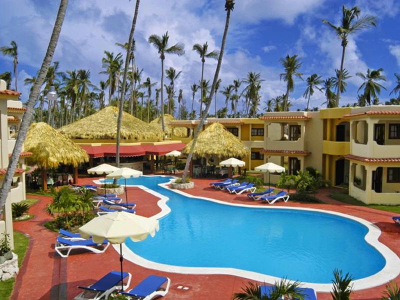 Hotel mit blauem Pool, Sonnenliegen, Palmen und gelben Gebäuden bei sonnigem Himmel.