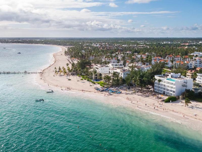 Strand mit klarem türkisfarbenem Wasser, weißen Sand, vielen Menschen und Gebäuden entlang der Küste.