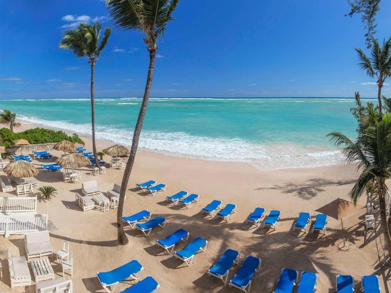 Strand mit blauen Liegestühlen, Palmen und türkisfarbenem Meer unter klarem blauem Himmel.