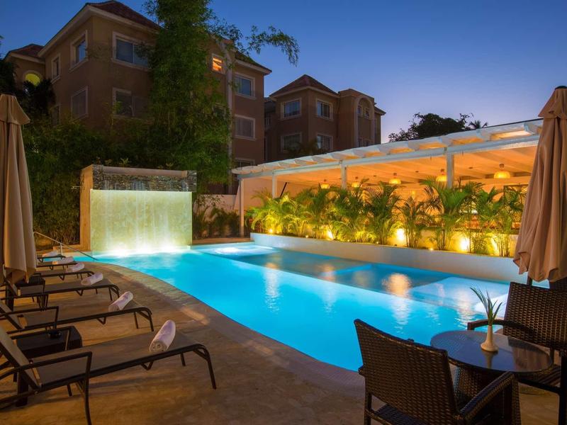 Beleuchteter Pool mit Wasserfall, Liegestühlen und Sitzbereich unter blauem Abendhimmel.
