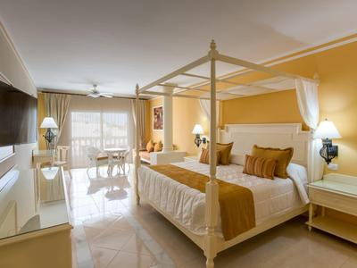 Bright hotel room with four-poster bed, armchairs, and balcony with sea view.