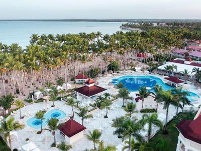 Vue d'une zone de piscine de complexe hôtelier avec des palmiers et la mer en arrière-plan sous un ciel nuageux.