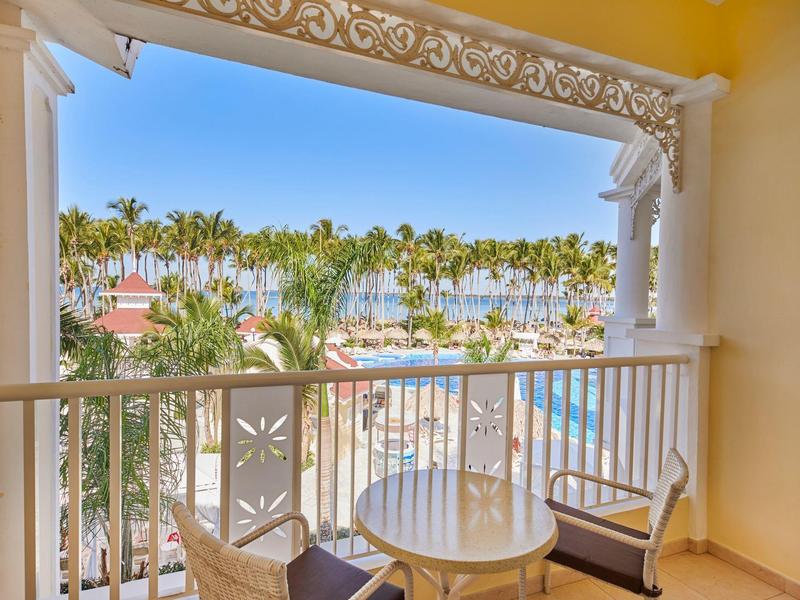 Balcony with table and chairs overlooking palm trees, buildings, and blue sky in a holiday resort.