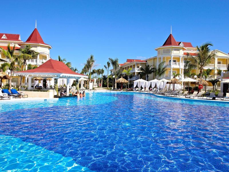 Large blue swimming pool at a resort with white umbrellas and red roofs under clear sky.