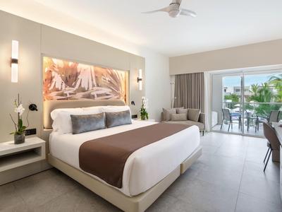 Modernes Hotelzimmer mit großem Bett, Schreibtisch und Balkon mit Blick auf Palmen.