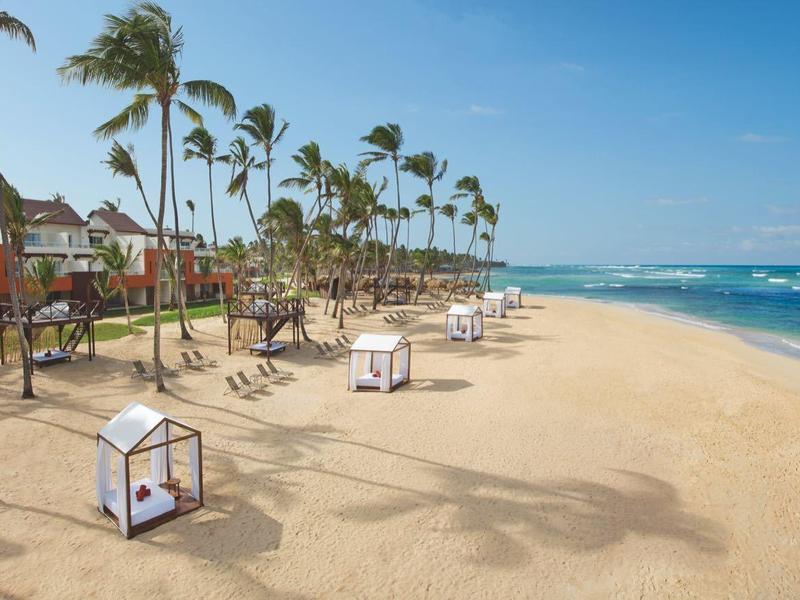 Weitläufiger Sandstrand mit Palmen, einer Reihe von Liegestühlen und blauer Meereskulisse unter klarem Himmel.