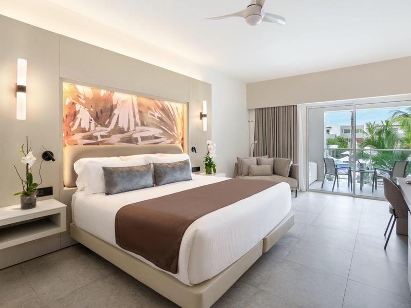 Modernes Hotelzimmer mit großem Bett, Schreibtisch und Balkon mit Blick auf Palmen.