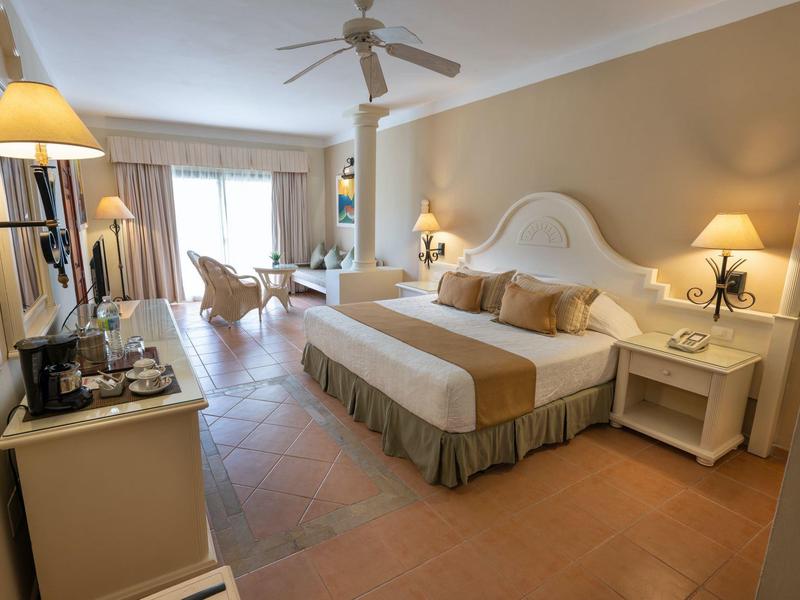 Habitación de hotel luminosa con cama grande, mesitas y zona de estar cerca de las puertas del balcón