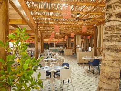 Offenes Restaurant mit Holzdach, weißen Tischen, blauen Stühlen und Pflanzen in warmem Licht.