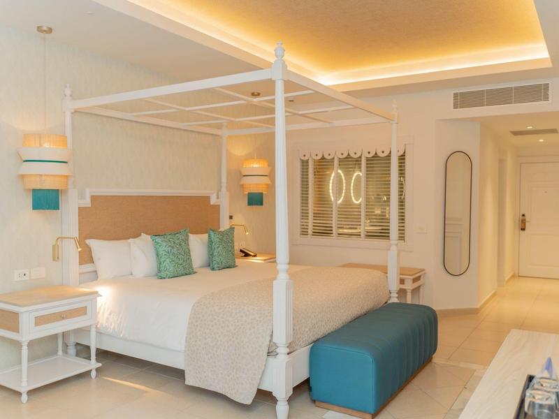 Helles Schlafzimmer mit weißem Himmelbett, beigem Bettwäsche und türkisfarbenen Kissen und Bank.