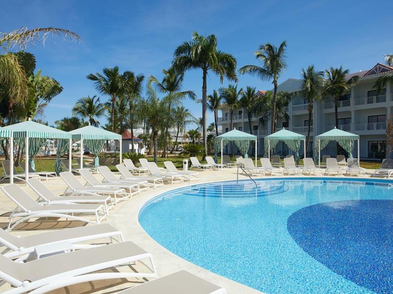 Großer, runder Pool mit blauer Wasserfläche, weißen Liegestühlen und grünen Pavillons unter Palmen.