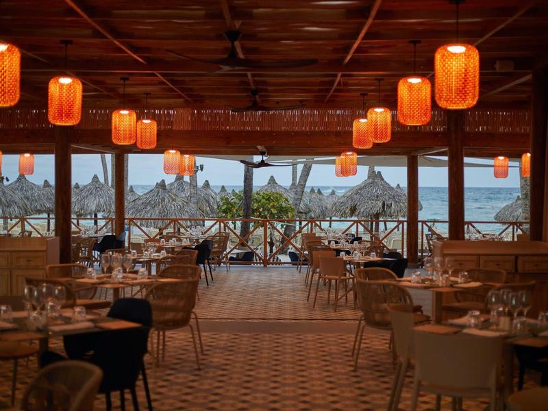 Offenes Restaurant mit Holzdecke, orangenen Lampen und Tischen mit Stühlen am Wasser.