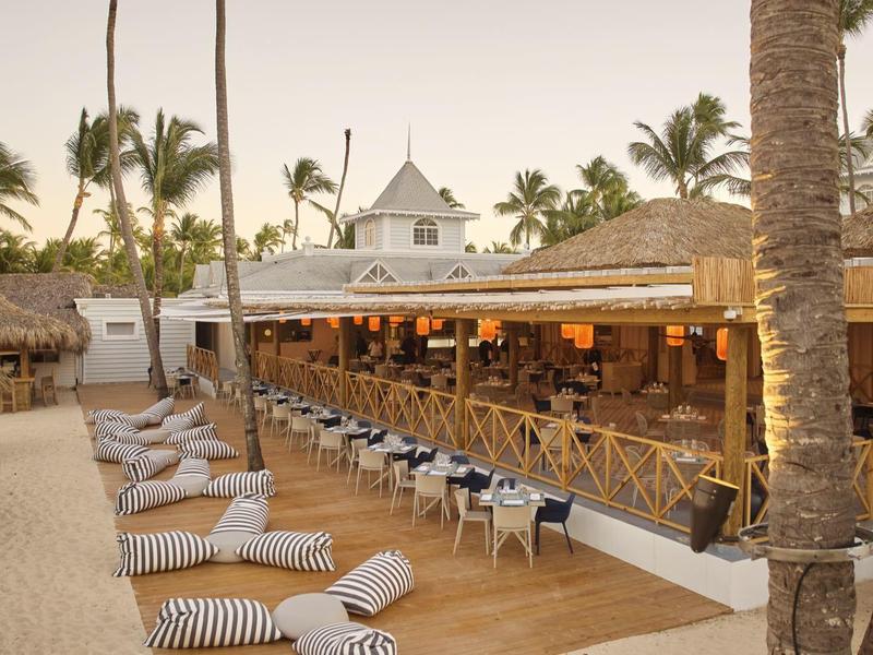 Strandbar mit Holzterrasse, Sitzgelegenheiten, Palmen und loungigen Kissen auf Sandfläche.
