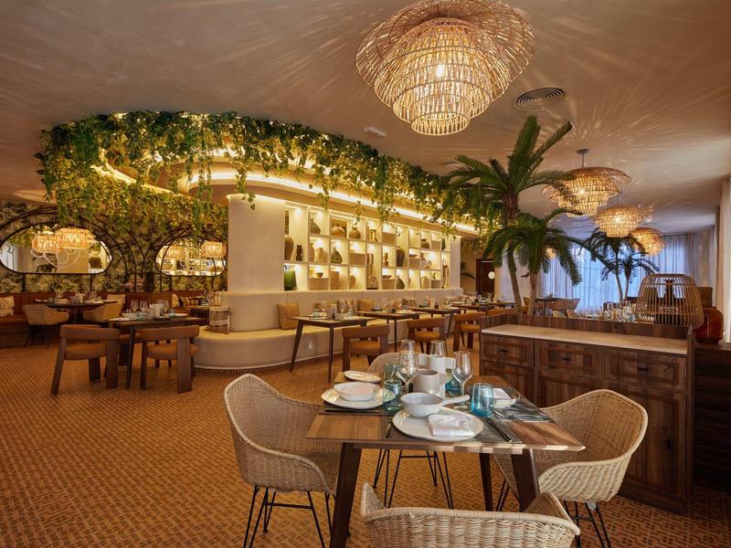 Gemütliches Restaurant mit Rattanmöbeln, grünen Pflanzen und warmem, goldenem Licht.