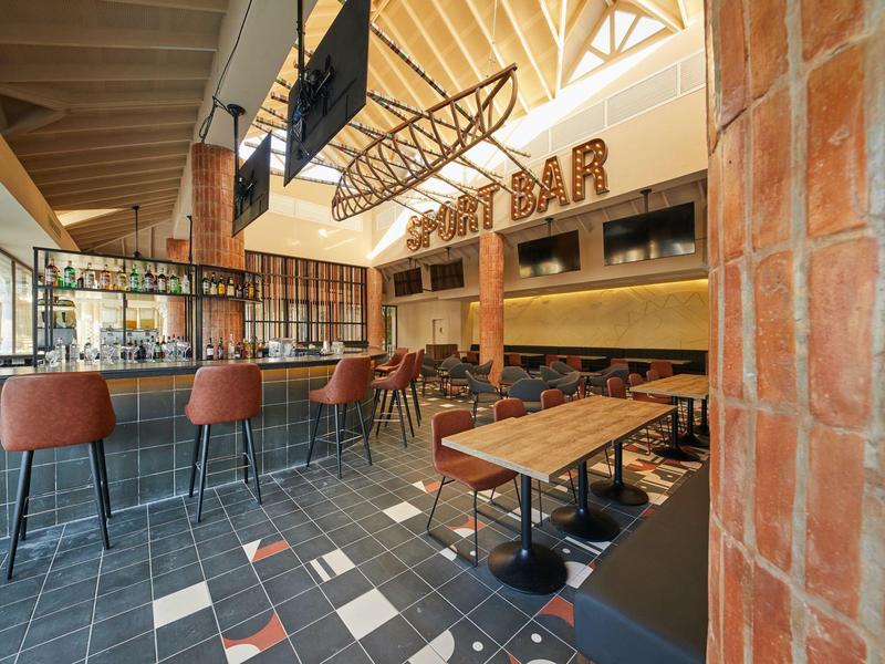 Modernes Barinterieur mit Holzdecke, roten Barhockern und langen Holztischen auf gefliestem Boden.