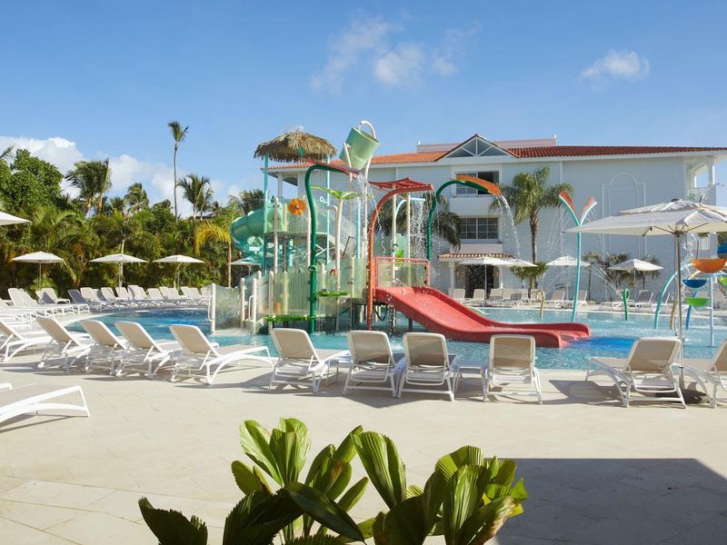 Freizeitbadbereich mit mehreren Liegestühlen, Wasserrutschen und Wasserspielen bei Sonnenschein.