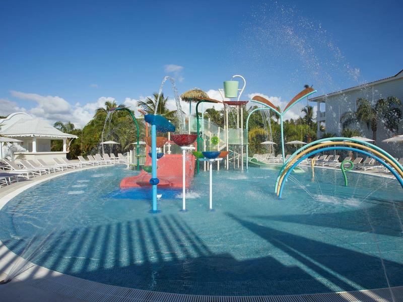 Kinderplanschbecken mit bunten Wasserspielen, Sonnenschirmen und weißem Hotelgebäude im Hintergrund.