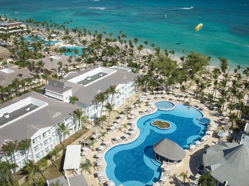 Großer Resortpool mit Palmen und weißen Gebäuden am türkisfarbenen Meerstrand.