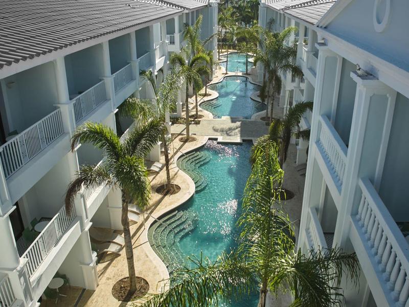 Blick auf einen langen, geschwungenen Pool zwischen zwei blauen Gebäuden mit Balkonen und Palmen.