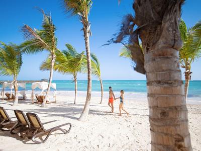 Coppia che passeggia su spiaggia di sabbia bianca con palme e lettini sotto un cielo azzurro.