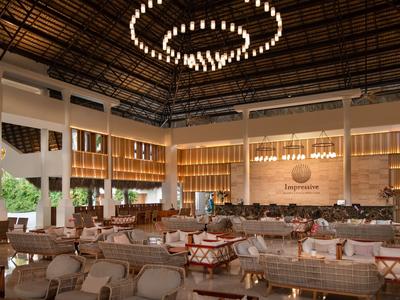 Przestronny hol hotelowy z wygodnymi fotelami, drewnianymi stołami i nowoczesnym żyrandolem pod wysokim sufitem.