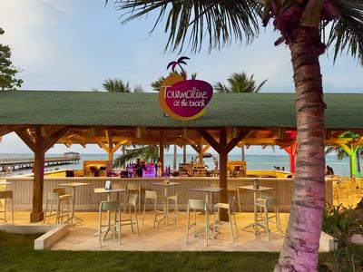 Bar sulla spiaggia con tetto in legno, sgabelli alti e palme sullo sfondo.