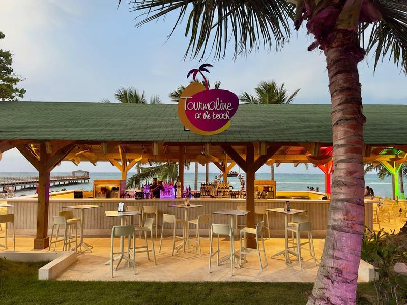 Bar sulla spiaggia con tetto in legno, sgabelli alti e palme sullo sfondo.