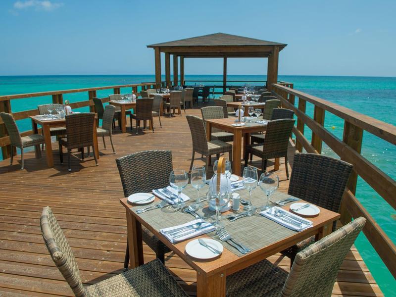 Area pranzo su un molo di legno sopra il mare azzurro cristallino con tavoli apparecchiati e sedie.