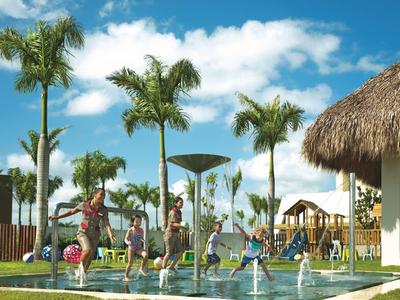 Kinder spielen mit Wasserfontänen in einem Pool vor Palmen und einem Haus mit Strohdach.