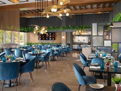 Modernes Restaurant mit blauen Sesseln, Holzboden, Hängepflanzen und großen Fenstern.