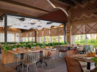 Helles Restaurant mit Holzmöbeln, Pflanzen und großen Fenstern, die viel Tageslicht hereinlassen.