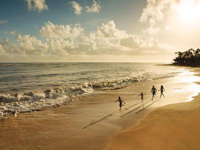 Strand mit sanften Wellen, vier Kindern beim Rennen, goldener Sand, bewölkter Himmel bei Sonnenuntergang.