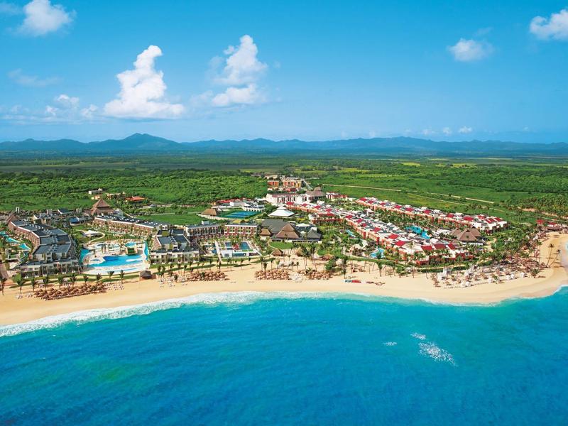 Große Küstenanlage mit Sandstrand, türkisblauem Wasser, grüner Landschaft und blauen Himmel.
