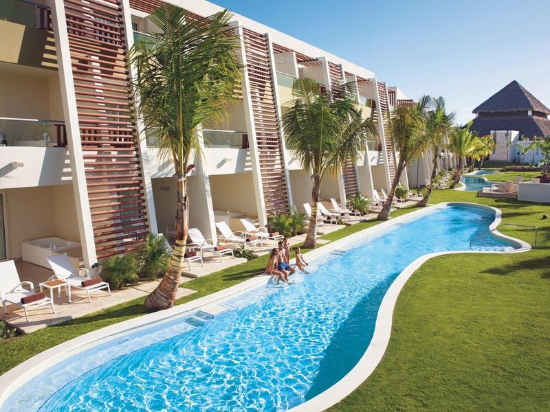 Langer, geschwungener Pool neben einem Hotel mit Liegestühlen und Palmen unter blauem Himmel.
