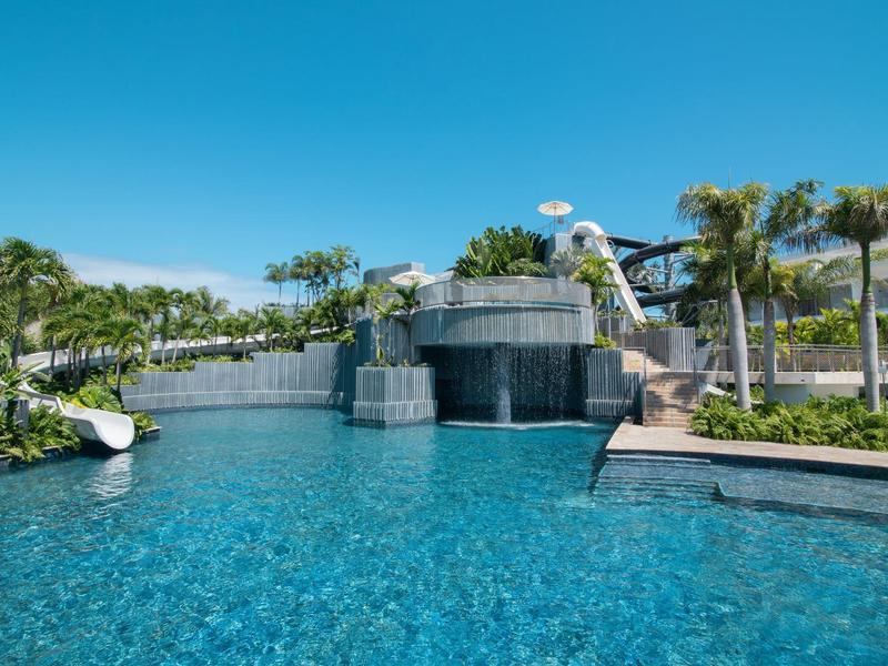 Großer Pool mit Wasserfall und umliegender moderner Hotelanlage unter klarem Himmel.