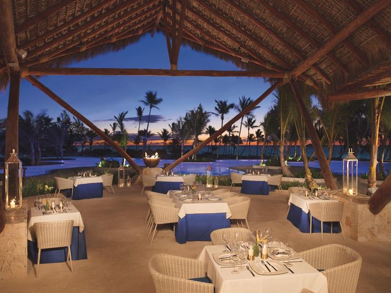 Avondterras van een restaurant met uitzicht op zwembad en palmboom onder een rieten dak.