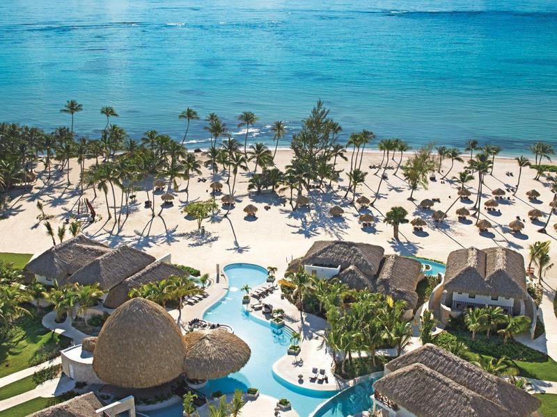 Uitzicht op een resort met zwembad, zandstrand en turquoise zee onder een blauwe lucht.