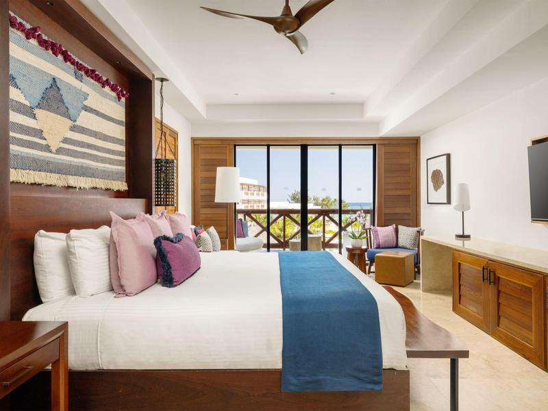 Modern hotelkamer met groot bed, kleurrijke kussens en uitzicht op balkon.