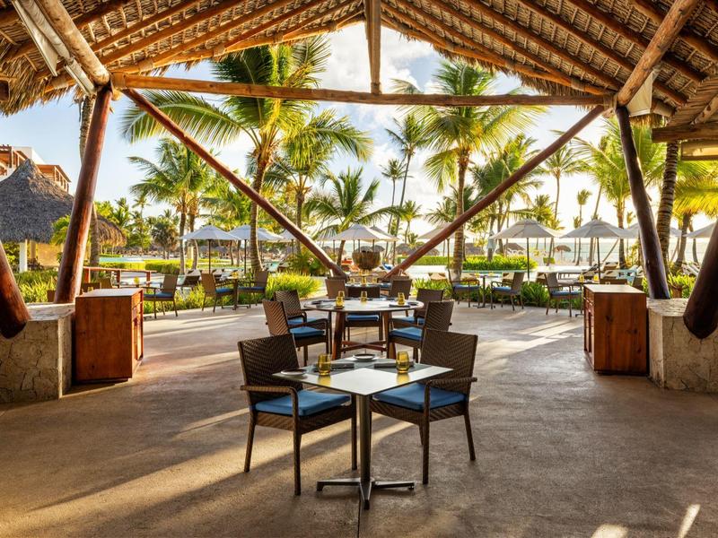 Open lounge met tafels en stoelen onder een rieten dak met uitzicht op palmbomen.