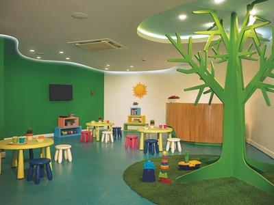 Kinderzimmer mit bunten Tischen, Stühlen und großem grünen Baum-Deko im Raum mit grünem Boden.
