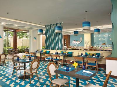 Modernes Restaurant mit blau-türkisem Farbkonzept, Holzstühlen, gemustertem Boden und großer Fensterfront.