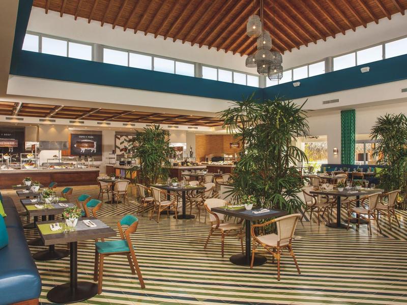 Modern hotel restaurant with seating, plants, and daylight from tall windows.